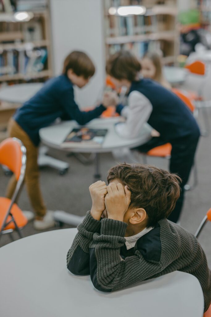 pexels-photo-7929440-7929440 A disheartened child sits alone in a school library while other kids interact.